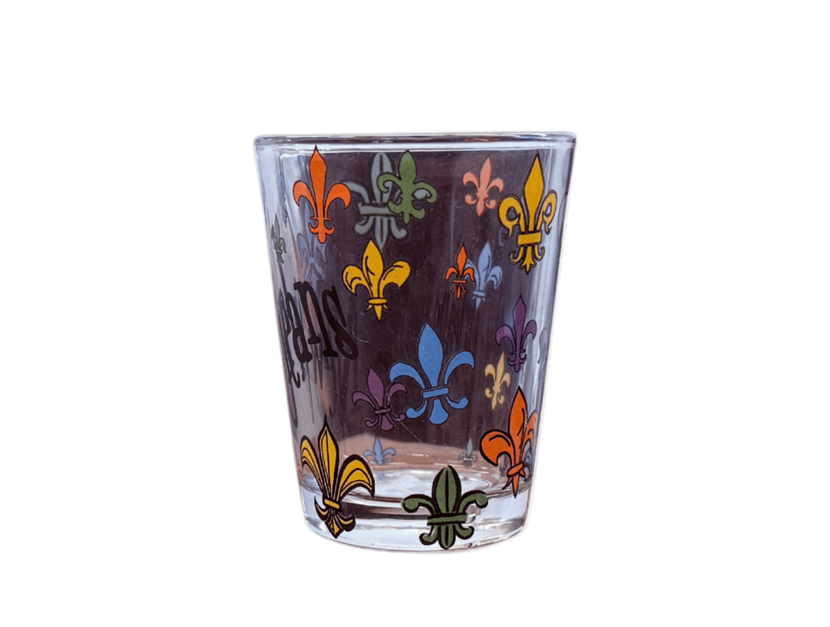 A clear glass shot glass decorated with colorful fleur-de-lis motifs in orange, yellow, blue, green, and purple on a dark background.