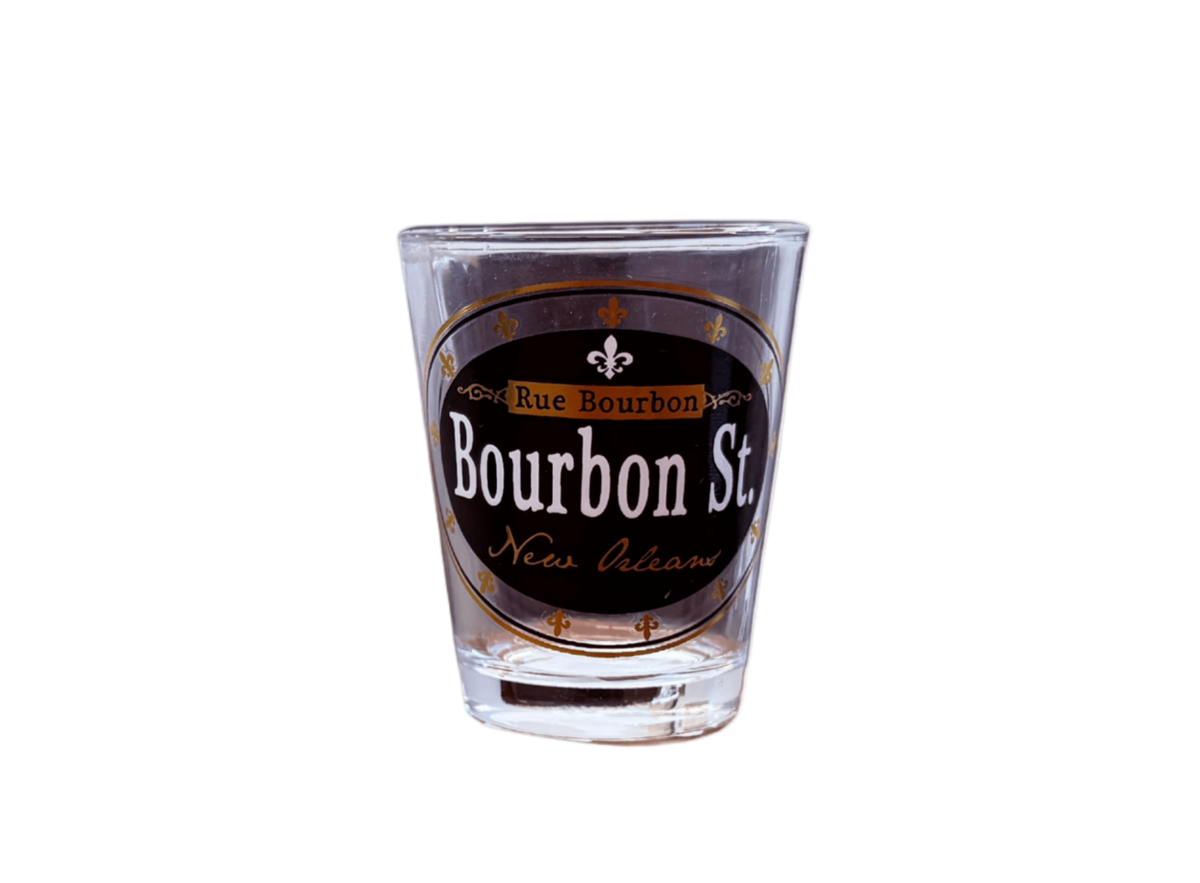 A clear glass shot glass featuring a black oval label with 'Bourbon St. New Orleans' and 'Rue Bourbon' in white and gold lettering, shown on a plain white background.