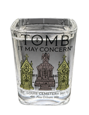 A clear glass shot glass featuring a black and white illustration of a cemetery monument with two green-tinted tombstones, and the text 'TOMB IT MAY CONCERN' and 'ST. LOUIS CEMETERY NO.