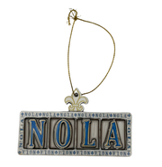 A rectangular metal Christmas ornament spelling 'NOLA' in raised letters with blue enamel fill, bordered by script text, hanging from a gold-tone chain on a plain white background.